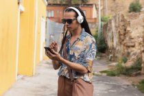 Frontansicht eines Mischlings mit langen Dreadlocks, der an einem sonnigen Tag in der Stadt unterwegs ist, mit Kopfhörern auf der Straße steht und ein Smartphone benutzt — Stockfoto