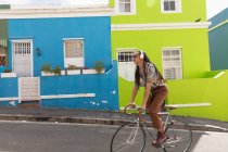 Seitenansicht eines gemischten Rennfahrers mit langen Dreadlocks, der an einem sonnigen Tag in der Stadt unterwegs ist, Kopfhörer trägt und sein Fahrrad auf der Straße fährt. — Stockfoto