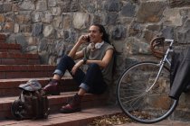 Вид сбоку мужчины смешанной расы с длинными ногами, который в солнечный день сидит на лестнице на улице и улыбается, используя смартфон, прислонившись на велосипеде к стене рядом с собой. — стоковое фото