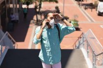 Вид спереди на смешанного расового человека с длинными дредами в городе в солнечный день, стоящего на улице в коронавирусной маске и использующего смартфон — стоковое фото