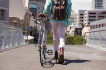 Vista posteriore bassa sezione dell'uomo con lunghi dreadlocks in giro per la città in una giornata di sole, indossando lo zaino, camminando per strada e ruote la sua bicicletta. — Foto stock