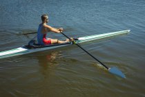 Висококутний вид на кавказький чоловічий веслувальник тренування і веслування на річці, тримаючи весла і сидячи в веслувальному човні в сонячний день. — стокове фото