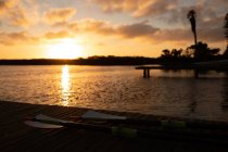Stunning landscape of the river by twilight, with a jetty, lying oars left after rowing training, with clear sky, clouds and sun reflecting in water — Stock Photo