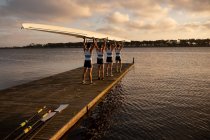 Rear view of a rowing team of four Caucasian men carrying a boat above their heads with arms raised, walking along a jetty on the river — Stock Photo
