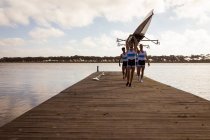 Front view of a rowing team of four Caucasian men carrying a boat above their heads with arms raised, walking along a jetty on the river — Stock Photo
