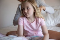 Vue de face gros plan d'une femme caucasienne profitant du temps en famille avec sa fille à la maison ensemble, la mère brossant les cheveux de sa fille assise sur un lit dans leur chambre — Photo de stock