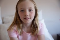 Retrato close-up de uma menina caucasiana feliz desfrutando de tempo livre em casa, sentado em uma cama em um quarto, sorrindo e olhando para a câmera, vestindo camiseta rosa — Fotografia de Stock