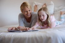Front view of a Caucasian woman enjoying family time with her daughter at home together, reading a book and lying on bed in their bedroom, smiling — Stock Photo