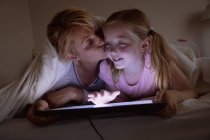 Вид спереди на кавказку, наслаждающуюся семейным отдыхом с дочерью дома вместе, лежащую на кровати в своей спальне, улыбающуюся и пользующуюся планшетным компьютером — стоковое фото