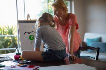На вигляд кавказька жінка, яка насолоджується сімейним часом зі своєю донькою вдома, сидить за столом у вітальні, малює і посміхається, дочка сидить на столі, малює на полотні. — стокове фото