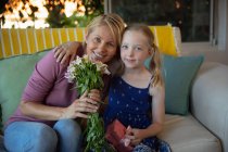 Портрет кавказької жінки, яка проводить сімейний час зі своєю дочкою вдома, сидячи на дивані у вітальні, тримаючи букет квітів, обіймаючи один одного, посміхаючись і дивлячись на камеру. — стокове фото