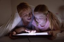 Vista frontal de uma mulher caucasiana desfrutando de tempo em família com sua filha em casa, deitada na cama em seu quarto à noite, sorrindo, usando tablet — Fotografia de Stock