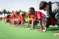 Side view of a multi-ethnic group of teenage male field hockey players training before a game, working out in a row, doing push ups on a hockey pitch on a sunny day — Stock Photo