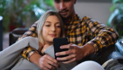 Передня сторона дивиться на молодого чоловіка змішаної раси і молоду кавказьку жінку, яка проводить час удома, сидячи в своїй вітальні, обіймаючи і використовуючи смартфон.. — стокове фото