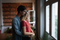 Бічний вид на молодого змішаного расового чоловіка і молоду кавказьку жінку, яка проводить час удома, стоїть біля вікна і обіймає одне одного.. — стокове фото
