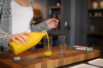 Frente vista seção meio da mulher de pé na cozinha preparando café da manhã, derramando suco de laranja para um copo . — Fotografia de Stock