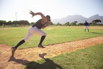 Vista lateral de un jugador de béisbol masculino de raza mixta, preparándose antes de un partido, ejercitándose, haciendo ejercicios, corriendo en un día soleado con su entrenador en el fondo - foto de stock