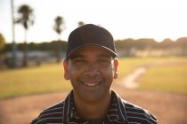 Ritratto di un arbitro di baseball maschile di razza mista, con indosso un'uniforme e un cappello, in piedi su un campo da baseball, che guarda una telecamera, sorridente — Foto stock