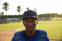 Porträt eines gemischten männlichen Baseballspielers, der eine Mannschaftsuniform, eine Mütze und Brustpolster trägt, auf einem Baseballfeld steht und in die Kamera blickt — Stockfoto