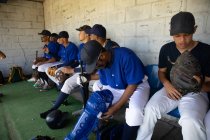 Vista lateral de una fila de jugadores de béisbol masculinos multiétnicos, preparándose antes de un juego, sentados en el vestuario, poniéndose almohadillas para las piernas, enfocándose mientras esperan, interactuando - foto de stock