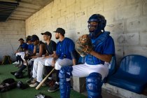 Vue latérale d'une rangée de joueurs de baseball multiethniques masculins, se préparant avant un match, assis dans le vestiaire, se concentrant pendant qu'ils attendent, interagissant — Photo de stock