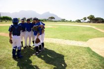 Side view of multi-ethnic team of male baseball players preparing before a game, in a huddle on a baseball field, listening to their captain giving them instructions, on a sunny day — Stock Photo