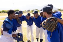Вид сбоку на многонациональную команду мужчин-бейсболистов, готовящихся перед игрой, в кучке на бейсбольном поле, слушающих, как их капитан дает им указания, в солнечный день — стоковое фото