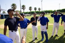 Вид сбоку на команду многонациональных бейсболистов-мужчин, готовящихся к игре, отдыхающих, питьевой воды и разговаривающих в солнечный день — стоковое фото