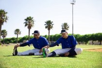 Vue latérale de deux joueurs de baseball de race mixte assis, étirant leurs jambes, lors d'une séance d'entraînement sur un terrain de jeu par une journée ensoleillée d'affilée, par une journée ensoleillée — Photo de stock