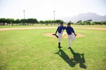 Side view of a Caucasian and a mixed race male baseball players leaning on each others shoulders, standing on one leg, holding and stretching the other leg, during a training session on a playing field on a sunny day — Stock Photo