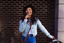 Ritratto di una felice donna di razza mista con lunghi capelli scuri in giro per le strade della città durante il giorno, indossando un cappello e una giacca di jeans, appoggiata sulla sua bicicletta sorridente alla macchina fotografica con muro sullo sfondo. — Foto stock