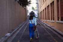 Vue arrière d'une heureuse femme métissée aux longs cheveux foncés dans les rues de la ville pendant la journée, portant un chapeau, un jean et une veste en denim, marchant avec son vélo avec des bâtiments en arrière-plan. — Photo de stock
