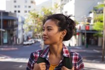 Side view close up of a happy mixed race woman with long dark hair out and about in the city streets during the day, carrying a backpack, wearing a checked shirt smiling with buildings in the background. — Stock Photo