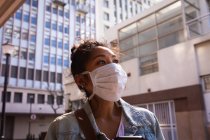 Low angle side view of a mixed race woman with long dark hair out and about in the city streets during the day, wearing a face mask against air pollution and coronavirus, standing with buildings in the background. — Stock Photo