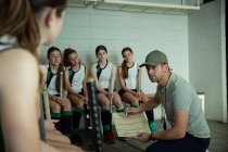 Side view of a Caucasian male field hockey coach interacting with a multi-ethnic group of female field hockey players, sitting in a changing room, showing them a game plan — Stock Photo