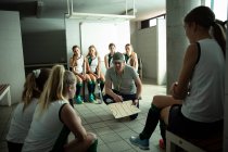Vorderseite eines kaukasischen männlichen Feldhockey-Trainers, der mit einer Gruppe kaukasischer Feldhockeyspielerinnen interagiert, die in einer Umkleidekabine sitzen und ihnen einen Spielplan zeigen — Stockfoto
