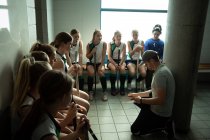 Side view of a Caucasian male field hockey coach interacting with a group of female Caucasian field hockey players, sitting in a changing room, showing them a game plan — Stock Photo