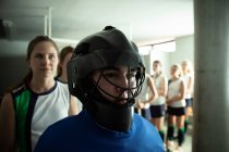 Side view close up of a Caucasian female field hockey players, preparing before a game, standing in a changing room, wearing a hockey helmet, with her teammates standing in a row behind her — Stock Photo