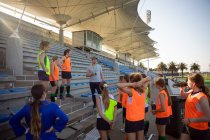 Vista lateral de um grupo de jogadoras de hóquei em campo caucasianas e seu treinador caucasiano treinando antes de um jogo, em pé em um estande, descansando, ouvindo seu treinador, em um dia ensolarado — Fotografia de Stock