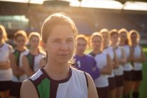 Porträt einer selbstbewussten kaukasischen Hockeyspielerin, die sich vor einem Spiel vorbereitet und an einem sonnigen Tag in die Kamera blickt, während ihre Teamkolleginnen in einer Reihe im Hintergrund stehen — Stockfoto