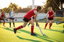 Side view of a Caucasian female field hockey player, during a field hockey game, running with a ball, holding a hockey stick, with her teammates and opponents in the background, on a sunny day — Stock Photo