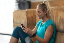 Seitenansicht einer athletischen kaukasischen Frau in Sportkleidung beim Crosstraining in einem Fitnessstudio, die eine Trainingspause im Sitzen einlegt und sich auf eine Box stützt, ein Smartphone benutzt und lächelt — Stockfoto