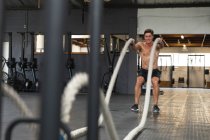 Vista frontal de um homem sem camisa, atlético caucasiano vestindo roupas esportivas cross training em um ginásio, exercitando-se com cordas de batalha — Fotografia de Stock