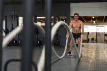 Vista frontal de um homem sem camisa, atlético caucasiano vestindo roupas esportivas cross training em um ginásio, exercitando-se com cordas de batalha — Fotografia de Stock