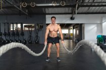 Vista frontale di un uomo caucasico atletico a torso nudo che indossa abiti sportivi, allenarsi in palestra, allenarsi con le corde da battaglia — Foto stock