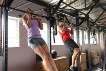 Side view of an athletic Caucasian man and woman wearing sports clothes cross training at a gym, holding on to a bar and doing chin ups beside each other — Stock Photo