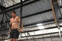 Frontansicht eines hemdlosen athletischen kaukasischen Mannes beim Crosstraining in einem Fitnessstudio, der sich an Turnringen hochschiebt und sein Körpergewicht hebt — Stockfoto