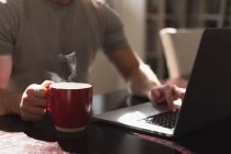 Vista lateral sección media del hombre pasar tiempo en casa, sentado junto al escritorio, utilizando su computadora portátil y sosteniendo una taza de bebida caliente. - foto de stock