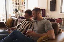 Vista lateral do casal masculino caucasiano relaxando em casa, sentado em um sofá, conversando e sorrindo, segurando xícaras de bebida enquanto usando um laptop juntos — Fotografia de Stock