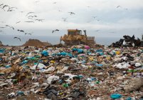 Troupeau d'oiseaux volant au-dessus de véhicules travaillant et déblayant des déchets empilés sur une décharge remplie de déchets avec un ciel nuageux et couvert en arrière-plan. Enjeu environnemental mondial de l'élimination des déchets. — Photo de stock